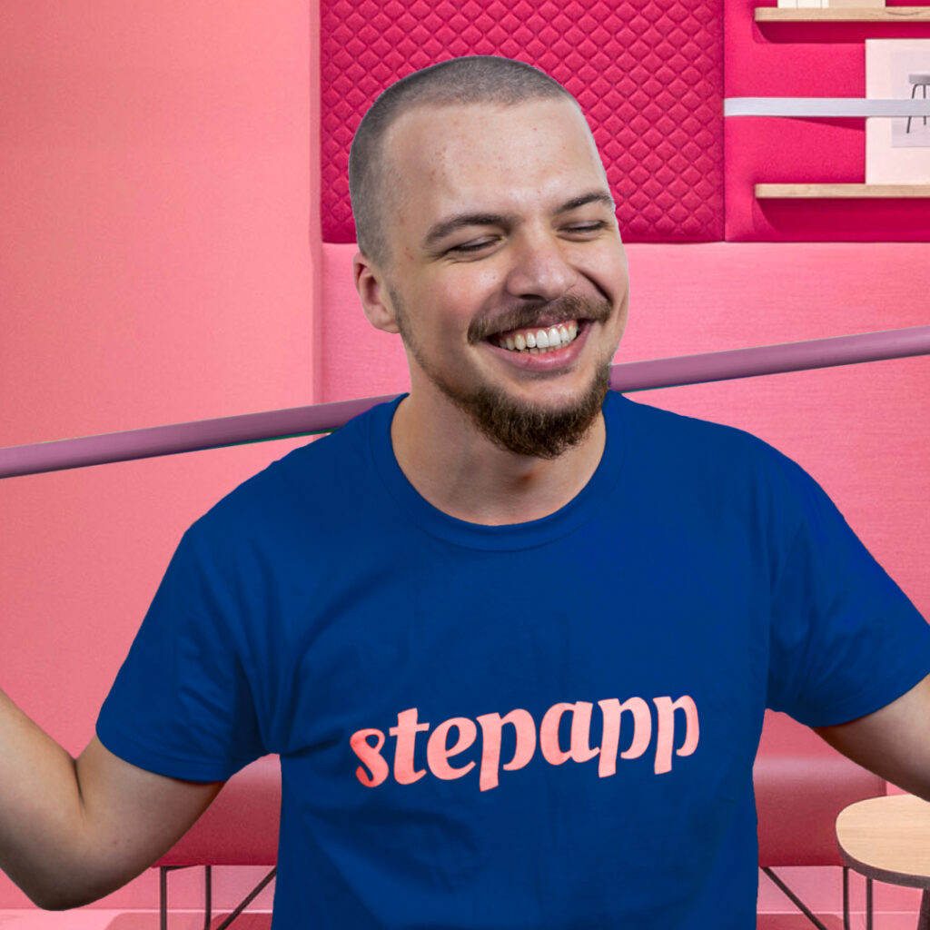 Mężczyzna z ogoloną głową i brodą, uśmiechnięty, z zamkniętymi oczami, nosi niebieski t-shirt z napisem „stepapp” napisanym różową czcionką. Stoi na różowym tle z pikowanymi panelami ściennymi i dobraną kolorystycznie półką, przywołując schludną estetykę często widoczną w usługach baza warszawa.