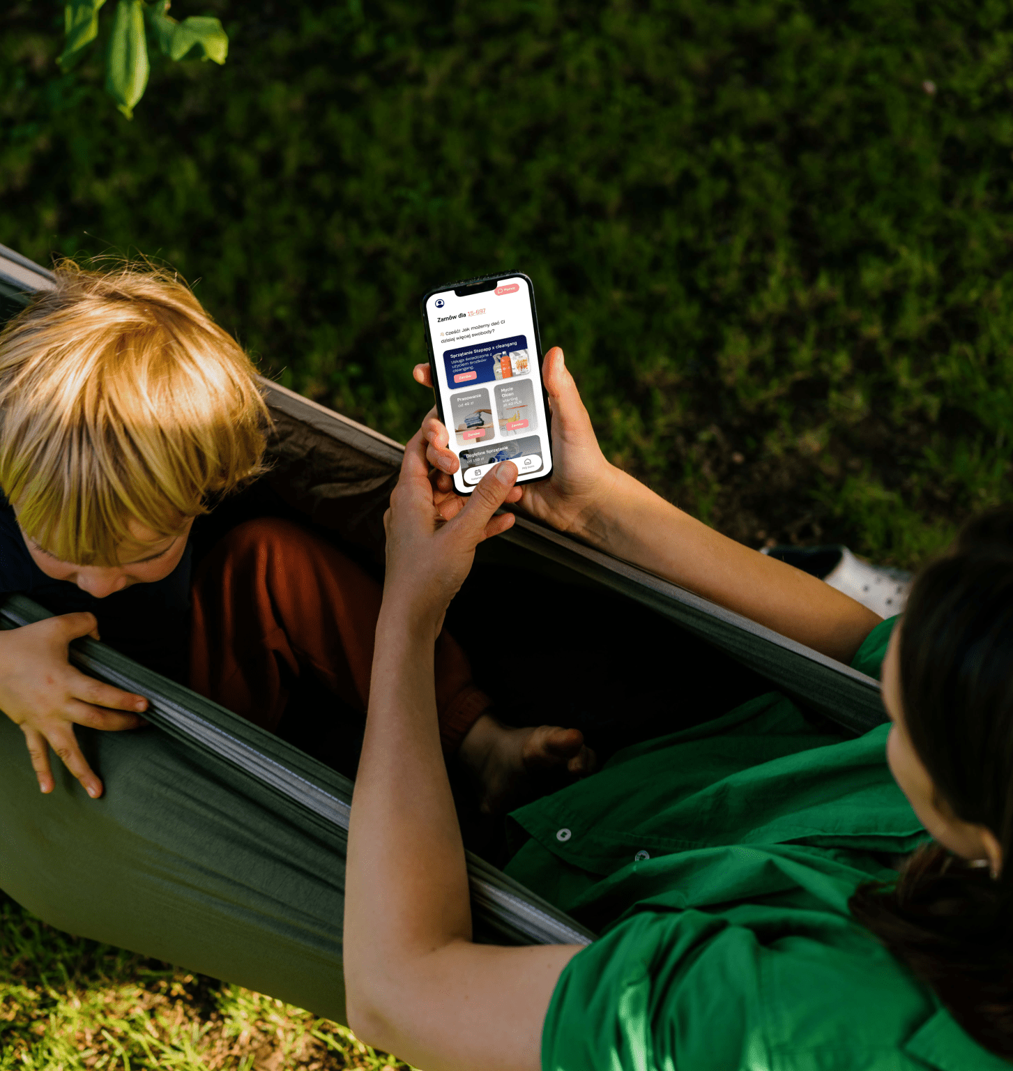 Osoba w zielonej koszuli siedzi w hamaku, trzymając smartfon, a obok niej dziecko. Na ekranie telefonu wyświetla się strona internetowa o oświetleniu domów. Scena rozgrywa się na zewnątrz, na tle trawy.