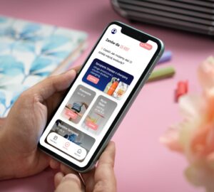 Osoba trzyma smartfon wyświetlający aplikację zakupową. Na ekranie wyświetlane są różne produkty gospodarstwa domowego, w tym środki czyszczące i ręczniki, z opcjami dodania ich do koszyka. W tle widać przedmiot z kwiatowym wzorem, długopis i kolorowe kwiaty — idealna scena dla osób zainteresowanych produktami mieszkań w Warszawie.